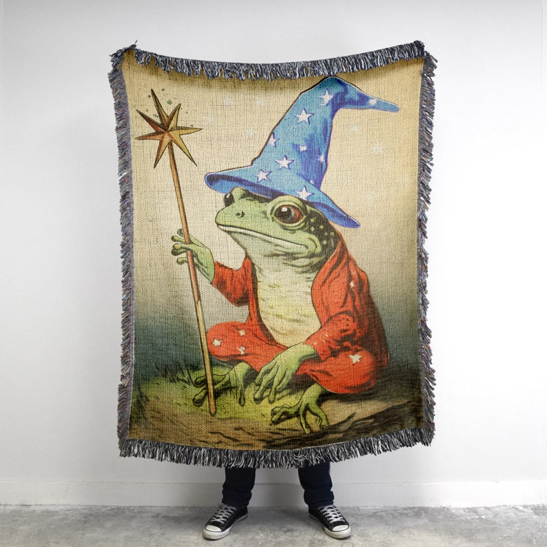 Op de afbeelding: Een geweven tapijtdeken met een cartoon kikker die een blauwe tovenaarshoed met witte sterren draagt. De kikker houdt een houten staf vast met een ster erop. De achtergrond is een bruin en groen landschap.