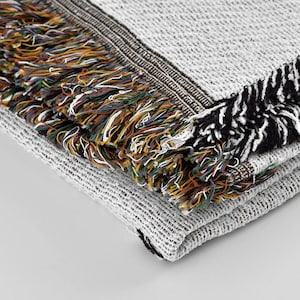 May include: A close-up of a gray and white woven throw blanket with fringe. The fringe is a mix of black, brown, green, and yellow threads.