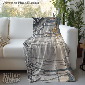 May include: A gray and white plush blanket with a printed design of trees and branches. The blanket is folded and draped over a white couch.