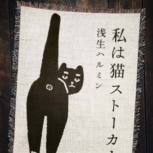 May include: A black cat with its tail raised high in the air, looking over its shoulder. The cat is on a cream-colored background with the text "I am a cat,  a stray" in Japanese.
