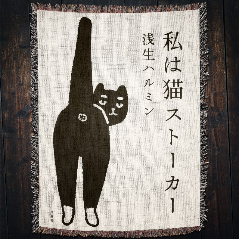 May include: A black and white woven tapestry featuring a cartoon cat with the Japanese text "I am a cat, born in a kiln" in black.