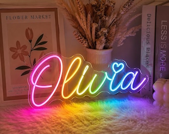 May include: A custom neon sign featuring the name Olivia in a cursive script font with a small heart icon above the letter i. The sign displays a vibrant rainbow gradient of pink, orange, yellow, green, and blue light, mounted on a clear acrylic backing.