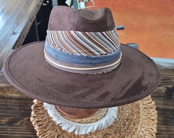 May include: A brown fedora-style hat with a wide brim. The hat features a striped band in shades of brown, blue, and cream. The brim is wide and the crown is indented. The hat is displayed on a woven surface.