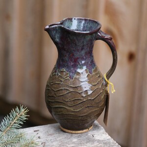 May include: A brown and purple ceramic pitcher with a textured surface and a handle. The pitcher is sitting on a wooden surface.