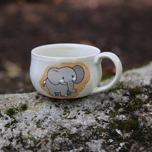May include: A white ceramic mug with a rounded shape and a single handle. The front of the mug features a hand-painted illustration of a small gray elephant with large ears and a trunk, set against a tan-colored oval background with white dots.
