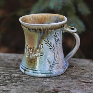 May include: A ceramic mug with a light blue, green, and brown glaze. The mug has a wide base and a handle. The word "Brew" is written on the side of the mug.