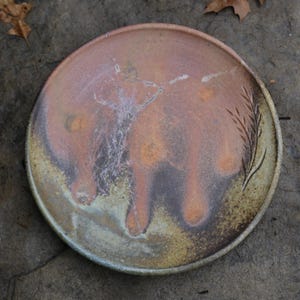 May include: A round ceramic plate with a unique color palette. The plate features a blend of orange, brown, and cream hues, with a textured surface. A decorative wheat stalk design is etched on the side. The plate is suitable for serving food.