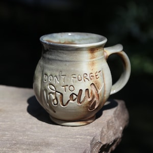 May include: A round, handmade ceramic mug with a light gray and tan glaze. The mug features a curved handle and the embossed text "DON'T FORGET TO PRAY" on the front. The surface has subtle brown and rust-colored accents throughout the clay body.