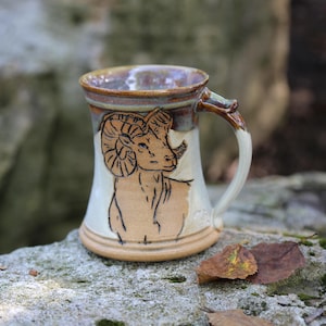 May include: A ceramic mug featuring a hand-drawn illustration of a bighorn sheep on the front. The mug has a flared base, a curved handle, and a two-tone glaze with a cream-colored body and a dark brown rim that extends down to the handle.