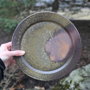 May include: A handmade ceramic plate with a speckled, earthy design. The plate features a dark brown rim and a center with shades of olive green and rust. The plate is held in a hand against a blurred outdoor background.