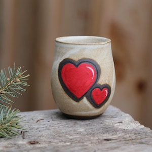 May include: A ceramic mug with a hand-painted design of two red hearts. The mug is a light beige color with a brown rim.
