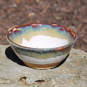 May include: A small ceramic bowl with a white interior and a multi-colored exterior featuring bands of cream, sage green, and a deep reddish-brown glaze around the rim. The bowl has a rounded shape and a smooth, glossy finish on the upper portion.