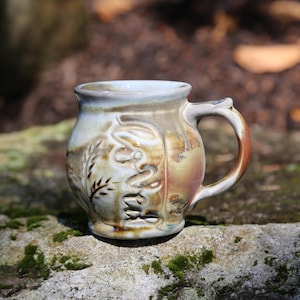 May include: A round ceramic mug with a glossy finish featuring a blend of cream, tan, and brown earth tones. The exterior surface displays embossed, swirling patterns and a stylized wheat stalk design, complemented by a curved handle on the right side.