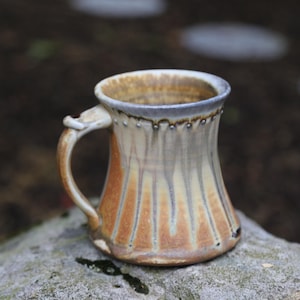 May include: A handmade ceramic mug with a unique design. The mug features a flared shape with vertical orange and tan stripes. The handle is curved and attached to the side. The rim is a darker color, and the mug is sitting on a rock.
