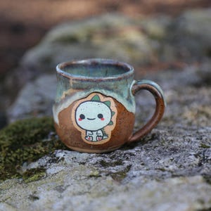 May include: A handmade ceramic mug with a dinosaur sticker. The mug has a brown base, a light green rim, and a brown handle. The dinosaur sticker is light green and white with a smiling face. The mug is sitting on a rock.