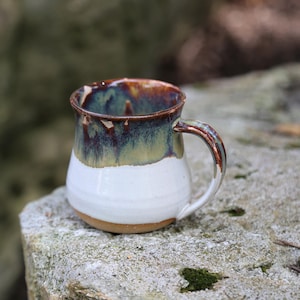 May include: A ceramic mug with a rounded body and a curved handle. The upper portion features a glossy glaze in shades of deep blue, green, and brown that drips downward, while the lower portion is finished in a smooth, solid white glaze.