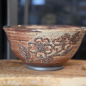 May include: A handmade ceramic bowl with a floral design. The bowl is brown with a light beige rim and a detailed embossed pattern of flowers and leaves. The bowl is approximately 6 inches in diameter.