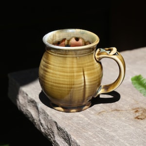 May include: A round ceramic mug with a glossy, variegated olive-green and brown glaze. The mug features a wide, bulbous body, a thick handle with a thumb rest, and a smooth, curved rim. The interior of the mug shows a reddish-brown clay base.