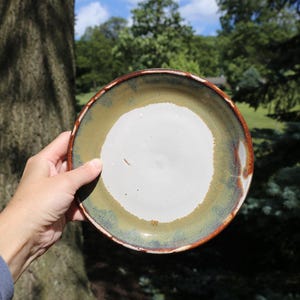 May include: A round ceramic plate featuring a smooth white center surrounded by a wide, mottled olive-green border. The outer rim of the plate is finished with a distinct, dark reddish-brown glaze that provides a clear contrast to the inner colors.