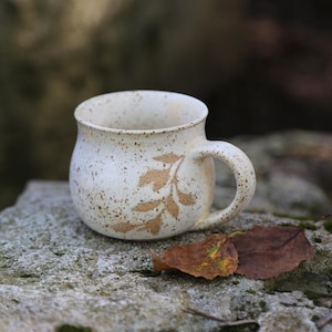 May include: A round ceramic mug with a speckled off-white glaze and a brown leaf pattern imprinted on the side. The mug features a curved handle and a wide, rounded body, showcasing a textured finish with small brown flecks across the surface.