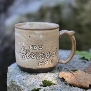 May include: A beige ceramic mug with a dark brown base and a curved handle. The front of the mug features the embossed white text "HOW blessed AM I" in a mix of block and script lettering. The surface has a speckled, textured appearance.