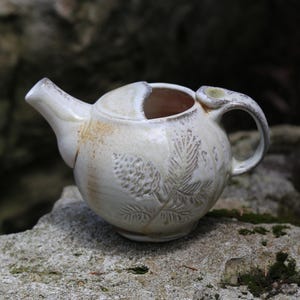 May include: A round, off-white ceramic teapot featuring a textured pinecone and needle relief pattern on its side. The teapot has a short spout, a curved handle, and an open top design with a smooth, glossy glaze finish and subtle tan accents.