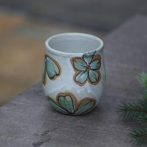 May include: A handmade ceramic tumbler with a light blue glaze. The tumbler is decorated with hand-painted green and brown floral designs. The cup is cylindrical with a slightly flared rim. The background is blurred, suggesting an outdoor setting.