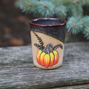 May include: A ceramic mug with a brown, black, and beige glaze. The mug features a hand-painted pumpkin with orange, yellow, and red accents.
