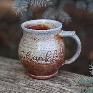 May include: A handmade ceramic mug with a brown and cream color scheme. The word "thankful" is etched into the side. The mug has a curved handle and is sitting on a wooden surface.