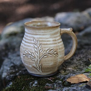May include: A ceramic mug with a rounded body and a curved handle, featuring a light beige and tan glaze with horizontal bands. The exterior surface is decorated with a carved, raised botanical pattern of a leafy stem on the front side.