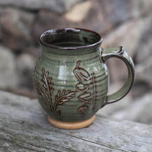 May include: A handmade ceramic mug in shades of green and brown, featuring embossed leaf and "Love" designs. The mug has a rounded shape, a sturdy handle, and a light brown base. The mug is approximately 4 inches tall.