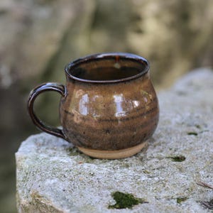 May include: A round ceramic mug with a glossy brown glaze featuring darker horizontal bands and a smooth, curved handle. The mug has a wide, bulbous body that tapers toward the rim, with a natural, unglazed tan base visible at the bottom.