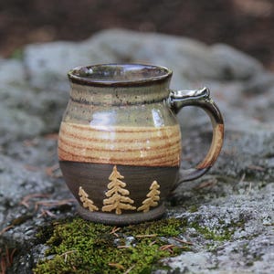 May include: A ceramic mug featuring a rounded body with a three-tone glaze. The top section is a mottled beige, the middle is a warm tan, and the bottom is a dark brown. Three embossed pine tree silhouettes are centered on the lower dark section.