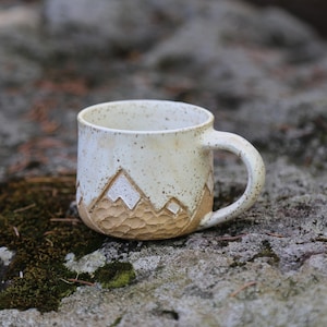 May include: A ceramic mug with a speckled off-white glaze on the upper portion and a textured, carved mountain range design in a tan earth tone on the lower portion. The mug features a rounded handle and a smooth, cylindrical body shape.