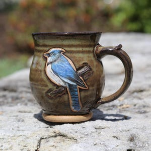 May include: A handmade ceramic mug with a detailed blue jay design. The mug is a mottled brown with a dark brown rim and handle. The blue jay is perched on a branch, with blue, white, and black coloring. The mug sits on a light brown base.