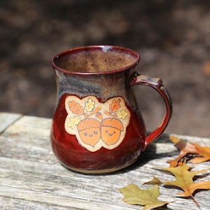 May include: A ceramic mug with a glossy deep red and dark brown glaze featuring a central decal of two smiling acorns with oak leaves. The mug has a rounded body and a sturdy handle, showcasing a smooth, reflective finish on the exterior surface.