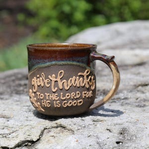 May include: A brown ceramic mug with a dark brown glazed rim and a handle. The mug features raised, embossed lettering that reads, "give thanks TO THE LORD FOR HE IS GOOD," along with a small embossed branch design on the lower left side.