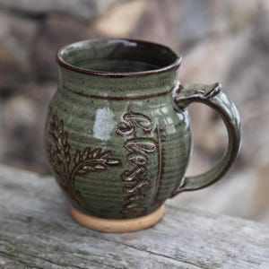 May include: A handmade ceramic mug in shades of green and brown. The mug features an embossed design with the word "Bless" and a leaf pattern. The handle is curved, and the mug sits on a small, light-colored base.