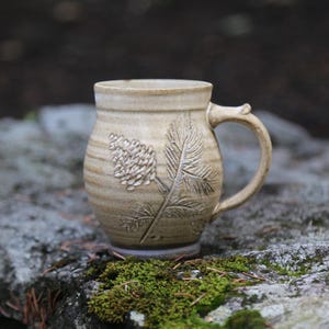 May include: A beige ceramic mug with a rounded body and a single handle. The exterior features a debossed botanical design of a pinecone and needle-like leaves. The surface has a smooth, glazed finish with subtle horizontal ridges around the base.