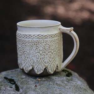 May include: A ceramic mug with a cream-colored glaze and a textured, raised lace-like pattern covering the exterior. The base of the mug is a dark brown, contrasting with the lighter upper section. It features a single handle on the right side.