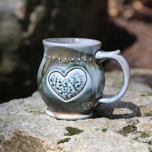 May include: A round ceramic mug with a glossy glaze in shades of blue, green, and brown. The mug features a prominent embossed heart shape on the front with a textured, dotted surface. A row of small raised dots encircles the upper rim area.