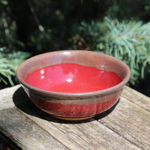 May include: A handmade ceramic bowl with a vibrant red interior and a brown exterior. The bowl is set on a weathered wooden surface, with a blurred green background of foliage. The bowl's rim has a darker, textured appearance.