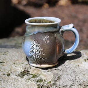May include: A ceramic mug featuring a rounded body with a glazed finish in shades of brown, blue, and cream. The exterior is decorated with an embossed pine tree and a crescent moon. It has a sturdy handle and a smooth, wide rim.