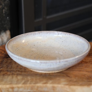 May include: A handmade ceramic bowl with a speckled, off-white interior and a slightly raised rim. The bowl is set on a wooden cutting board, showcasing its rustic charm. The bowl's exterior has a light brown rim.