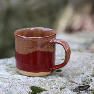 May include: A ceramic mug featuring a two-tone glaze design. The upper portion of the mug has a speckled tan and brown finish, while the lower section is a solid, deep red. It includes a curved handle on the side and a smooth, rounded rim.
