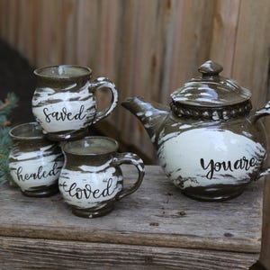 May include: A ceramic tea set with a teapot and three mugs. The set is brown and white with black lettering that says "Saved", "Healed", "Loved", and "You are". The teapot has a lid and a curved handle.
