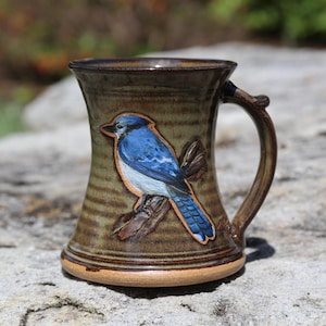 May include: Handmade ceramic mug with a detailed blue jay illustration. The mug features a brown handle and a textured, earthy-toned body. The bird is perched on a branch, adding a touch of nature-inspired art to the design.