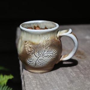 May include: A ceramic mug with a rounded body and a handle, featuring a cream-colored glaze on the upper portion and a natural brown finish on the lower section. The exterior is embossed with a detailed pine cone and needle branch design.