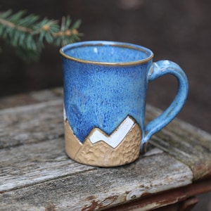 May include: A blue ceramic mug with a white and brown mountain design. The mug has a handle and is sitting on a wooden surface.
