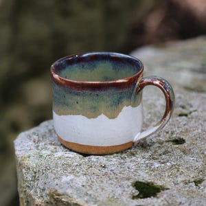 May include: A ceramic mug featuring a rounded body and a curved handle. The upper portion of the mug is glazed in a deep green and brown tone, while the lower portion is finished in a matte white glaze, revealing a natural tan clay base at the bottom.